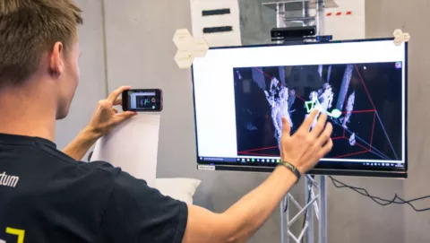 Mann steht mit Handy vor einem Bildschirm und ist mit der Hand und einem Kabel verbunden. Visualisierung der eignen Bewegung.