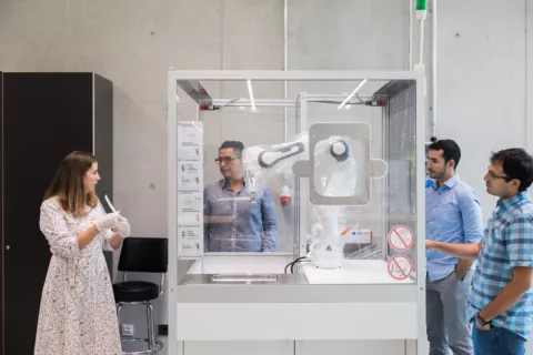 Vier Menschen stehen um eine gläserne Box. Darin ein Roboterarm, der durch Plastikfolie geschützt wird.