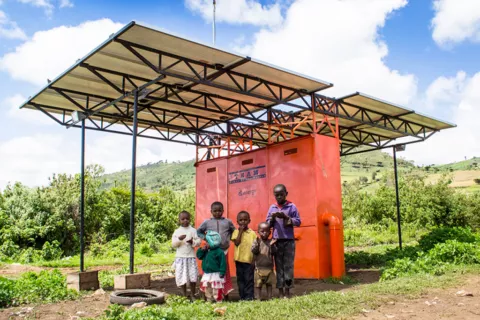 Beispiel-Foto eines Mini-Grid. Solarflächen an einer großen roten Box. Darunter Menschen im Schatten.