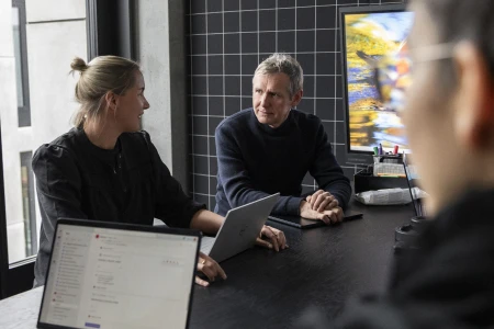 Two people sit at a table in a meeting room talking while a third person is blurred in the foreground.