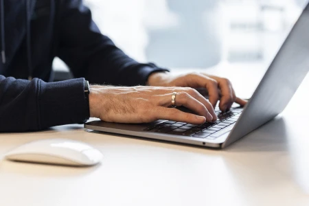 Hände tippen auf der Tastatur eines Laptops auf einem Tisch mit einer Computermaus daneben.
